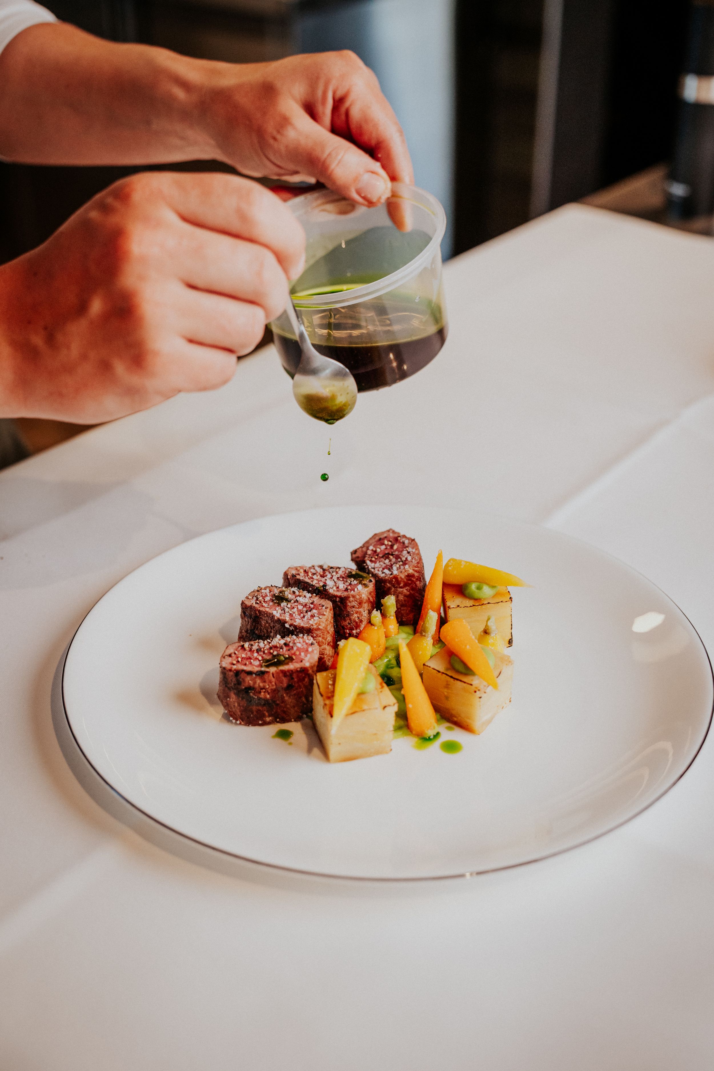 Chef drizzling sauce over plated meat and vegetables on white dish.
