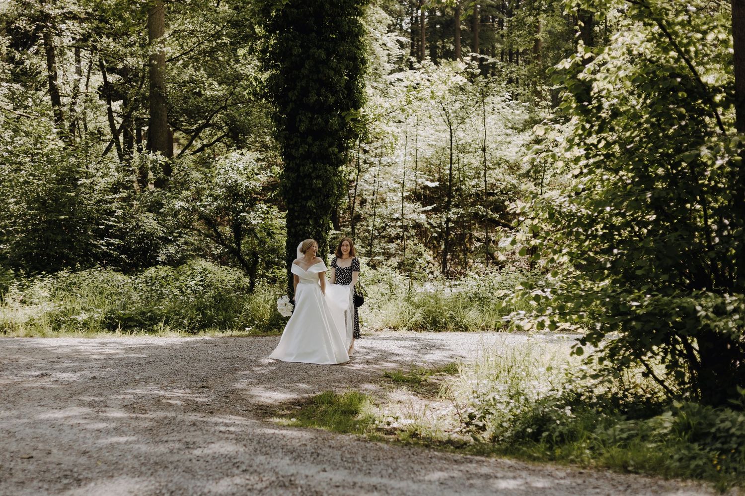 A wedding couple walks together on a sunlit forest path surrounded by lush greenery.