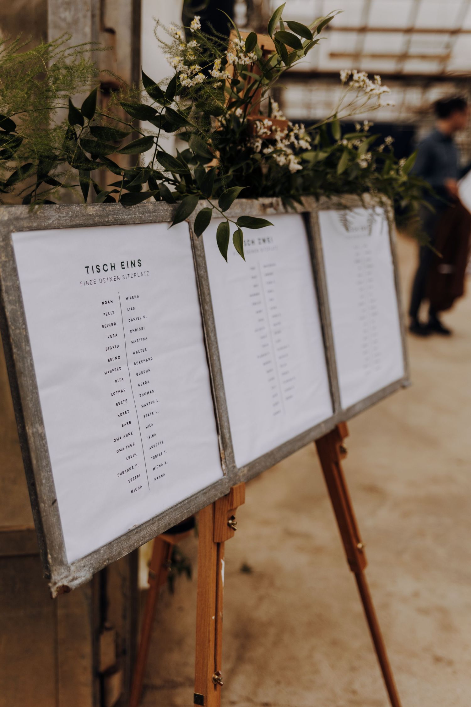 Wedding seating chart displays with greenery decorations mounted on wooden easels.