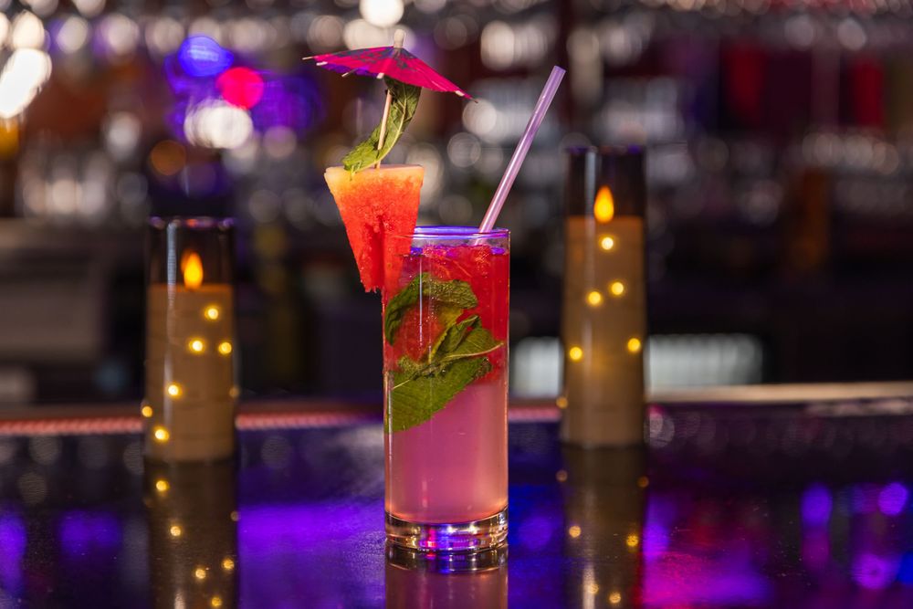 Colorful cocktails with garnishes sit on a dimly lit bar counter with glowing lights in the background.
