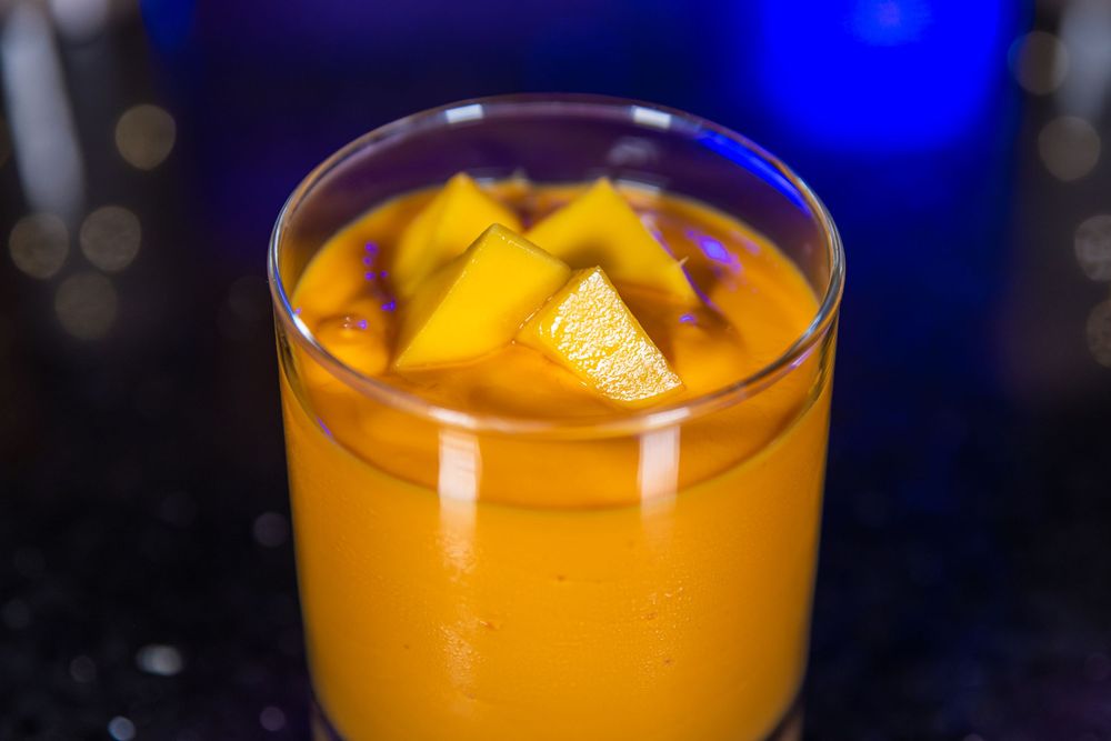 A vibrant orange mango smoothie garnished with fresh mango chunks in a clear glass against a dark background.