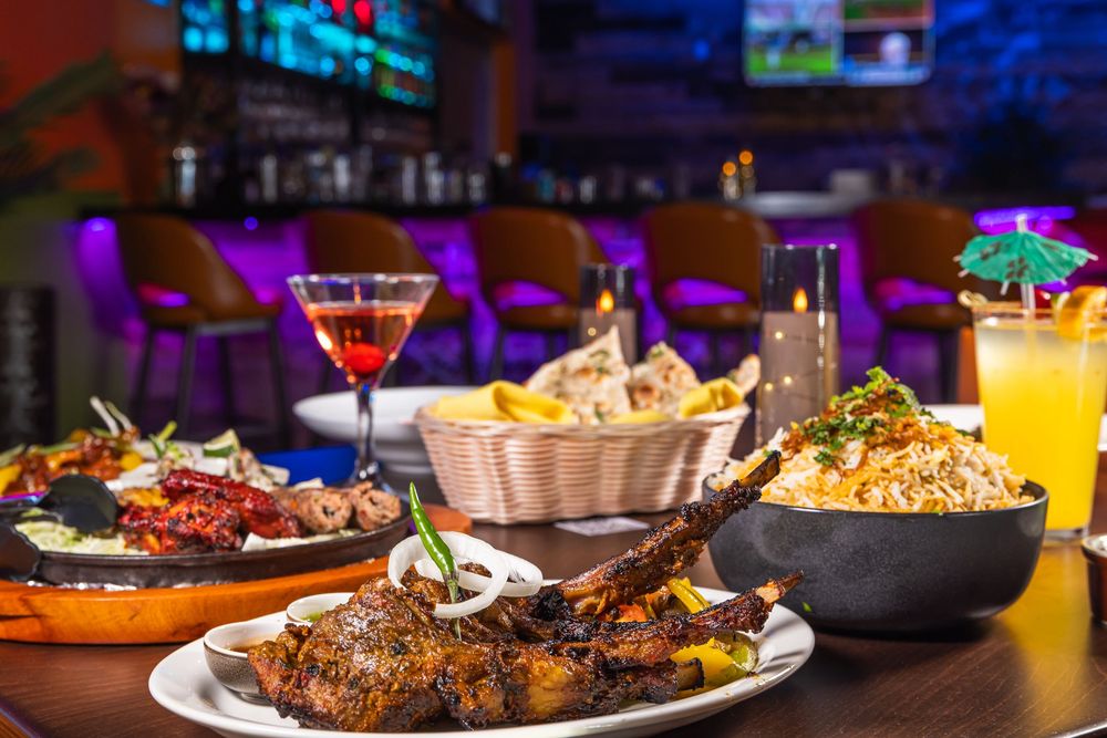 Colorful nightclub bar scene with cocktails, plates of food, and ambient blue lighting creating an upscale dining atmosphere.