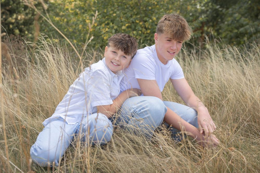 Two young boys in white shirts and jeans explore together in a field of tall grass on a sunny day.