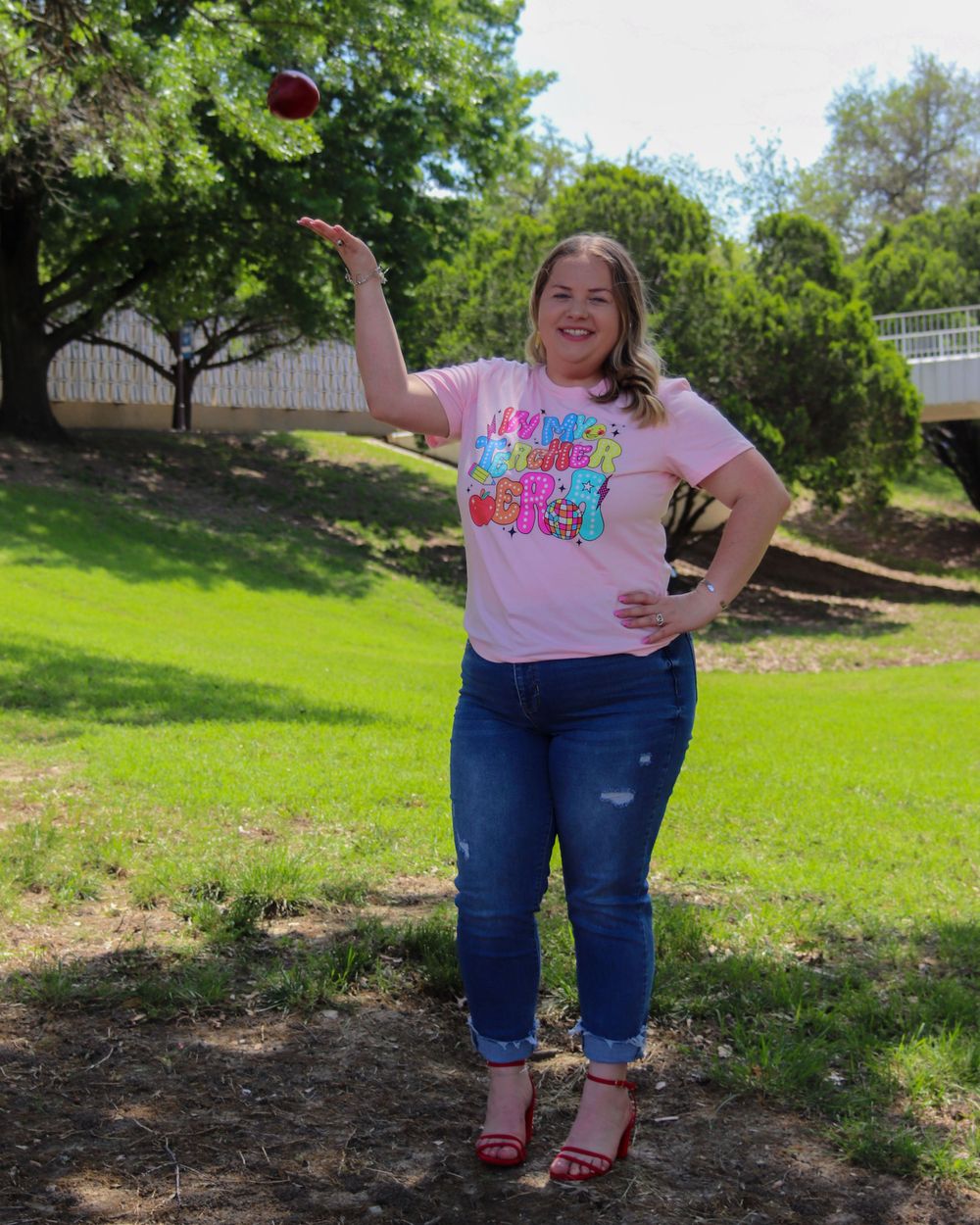 A person tosses a red object while posing on grass under trees wearing a pink t-shirt and distressed jeans with red shoes.