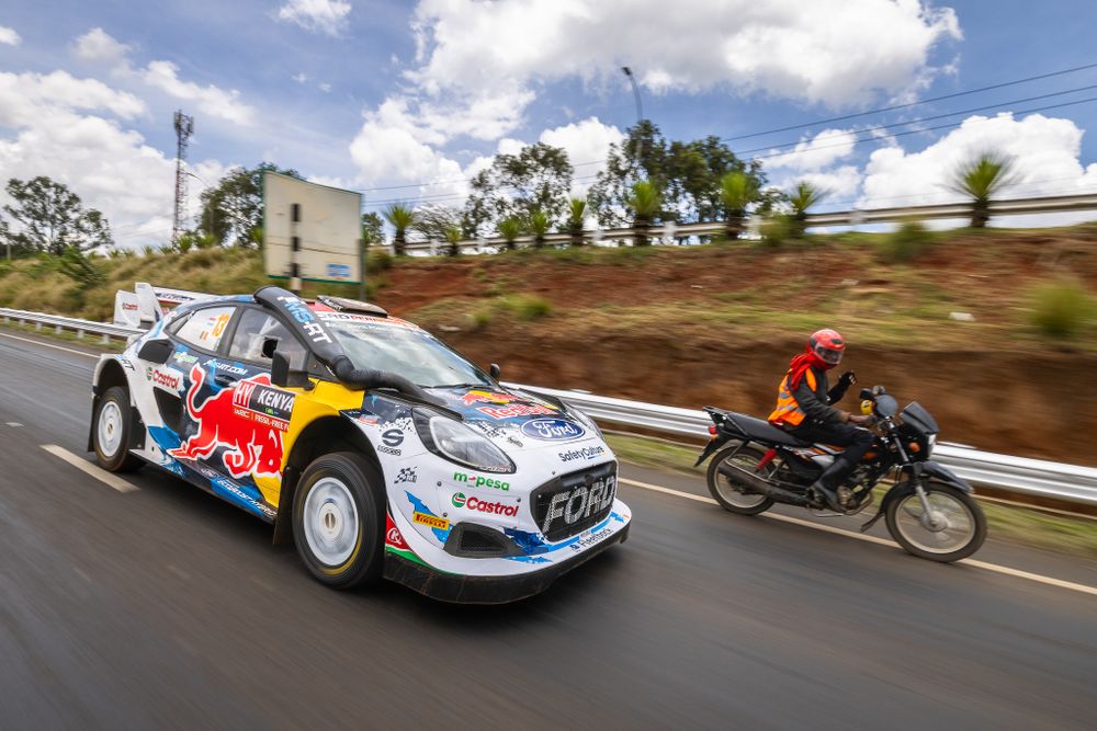 Red Bull rally car speeds past motorcycle on paved mountain road.
