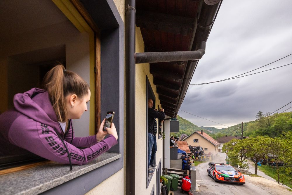 Rally car passes below spectator window during mountain stage race.