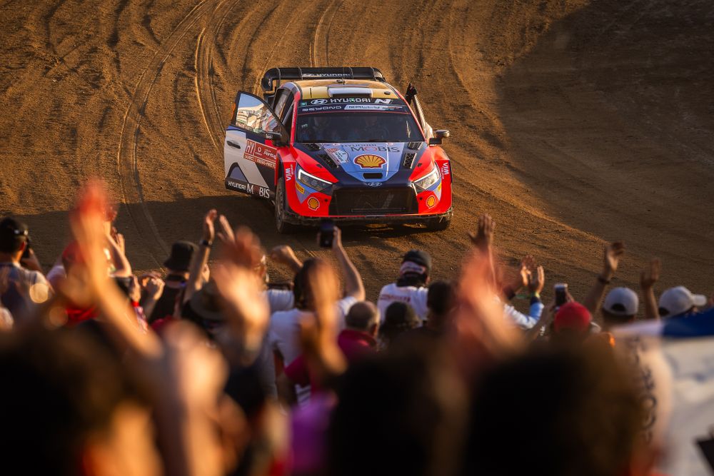 Rally car races past excited crowd on dirt track with dramatic lighting.
