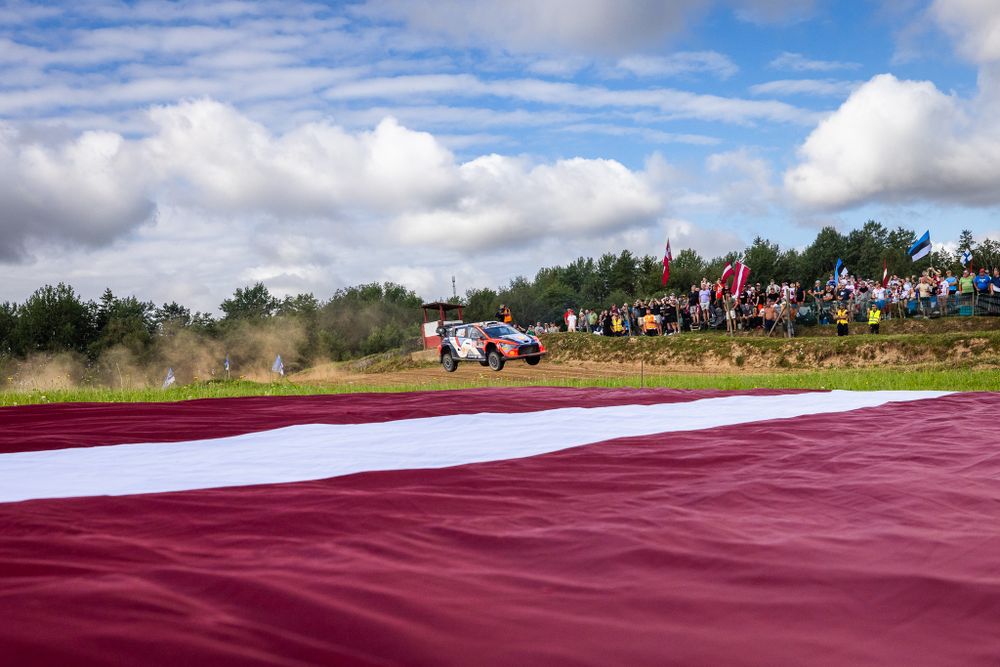 Rally car racing past large red and white flag during competition.