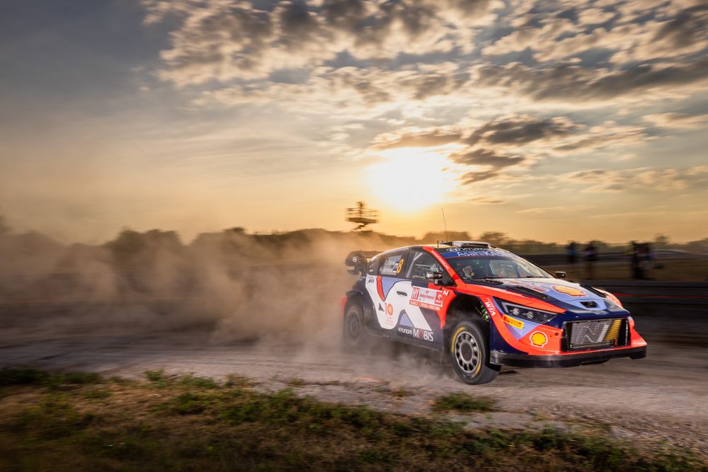 Rally car kicking up dust against dramatic sunset sky during off-road competition.