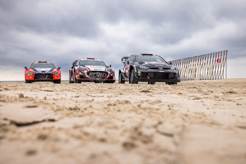 Three rally cars lined up on sandy beach under cloudy sky.