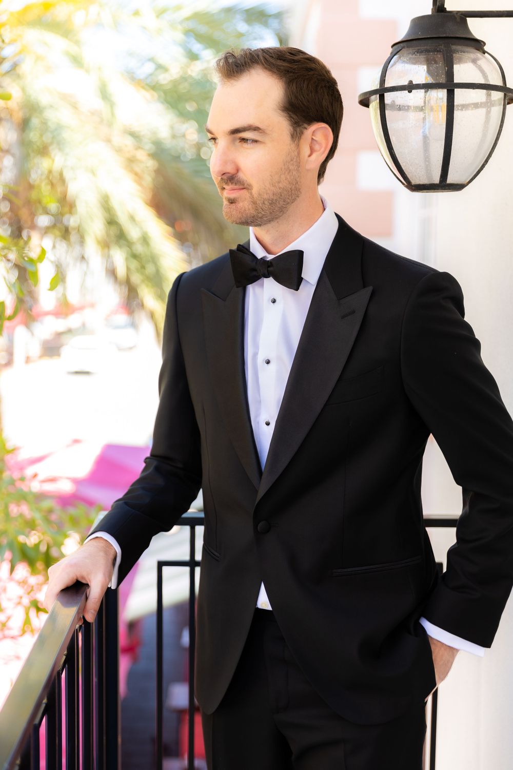Groom in classic black tuxedo posing against white pillars outdoors.