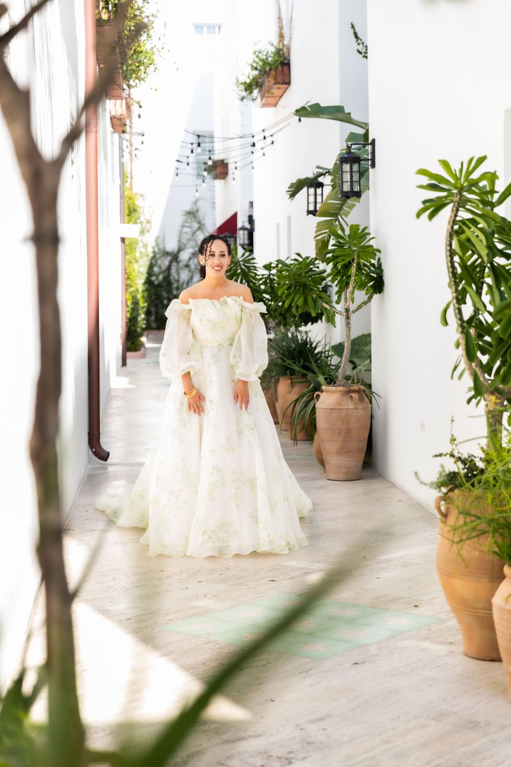 Bride in flowing white gown walks through Mediterranean courtyard with potted plants.