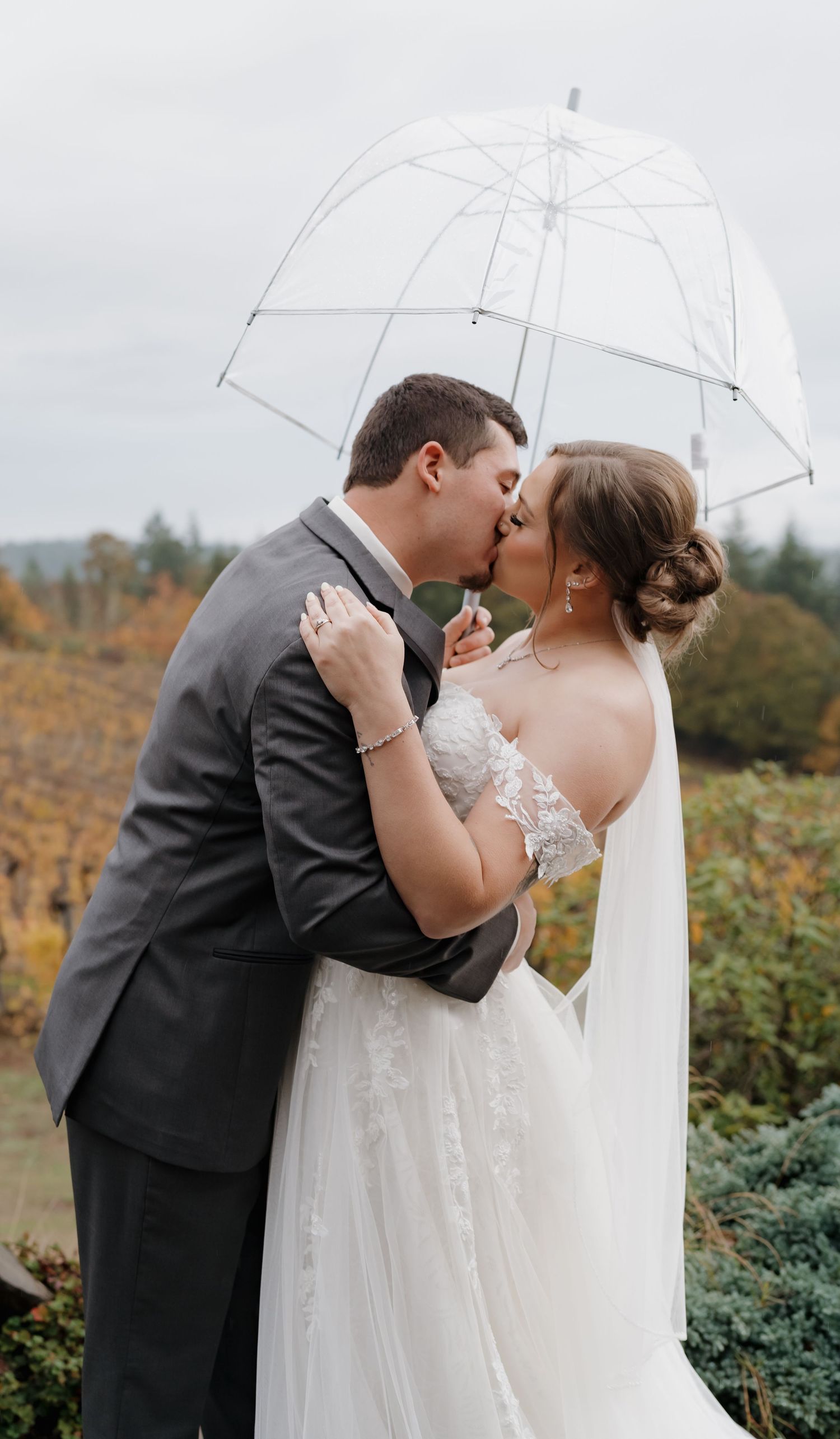 A romantic wedding couple shares a kiss under a clear umbrella against an autumn backdrop with yellow foliage.