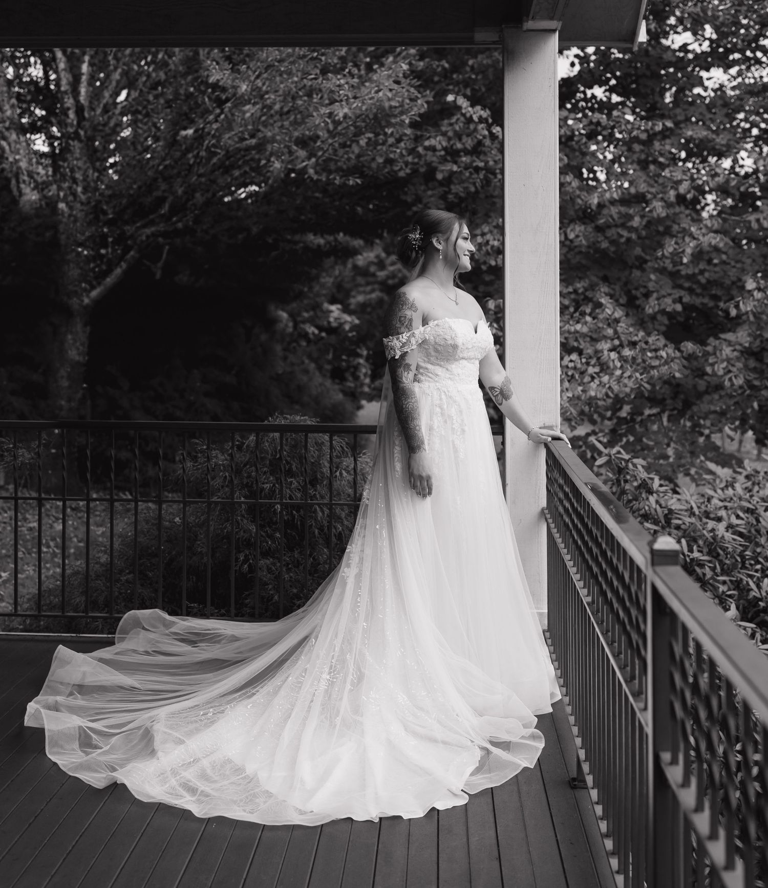 Black and white bridal portrait showing flowing wedding dress with long train on outdoor wooden deck with iron railing.