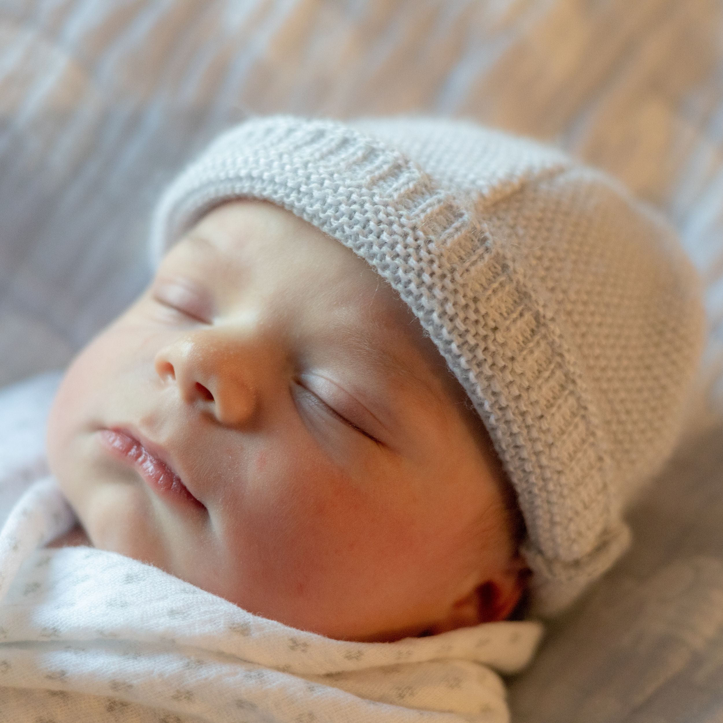 Sleeping baby wearing a soft knitted gray beanie hat rests peacefully against white bedding.