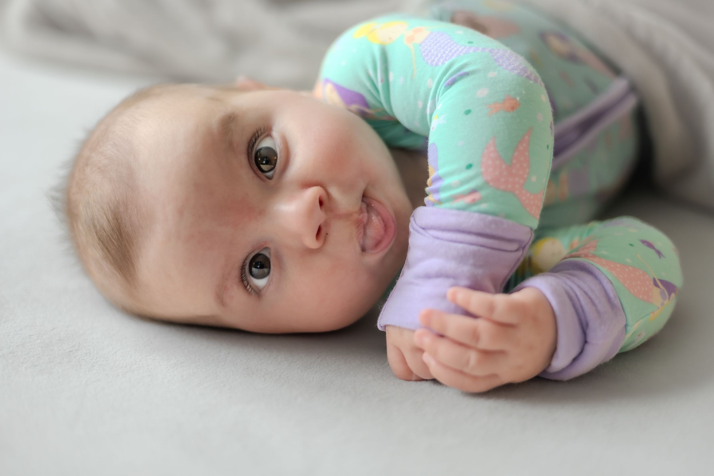 Baby wearing mint green and purple sleeper outfit lays on grey bedding.
