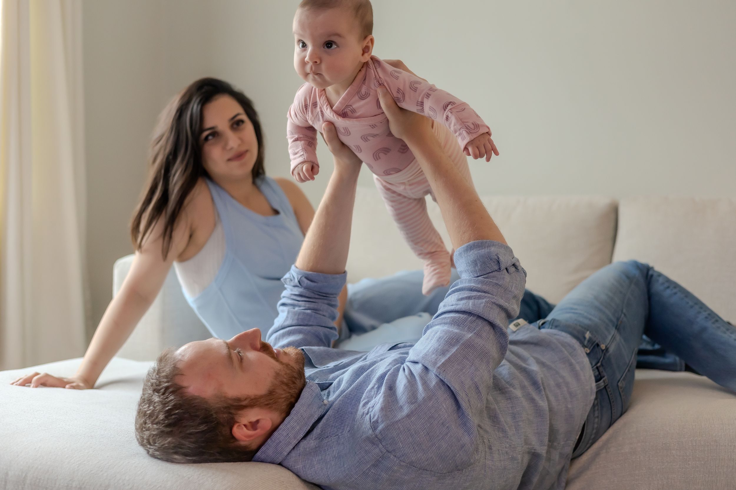 A parent lifts a baby wearing pink while relaxing on a couch at home.