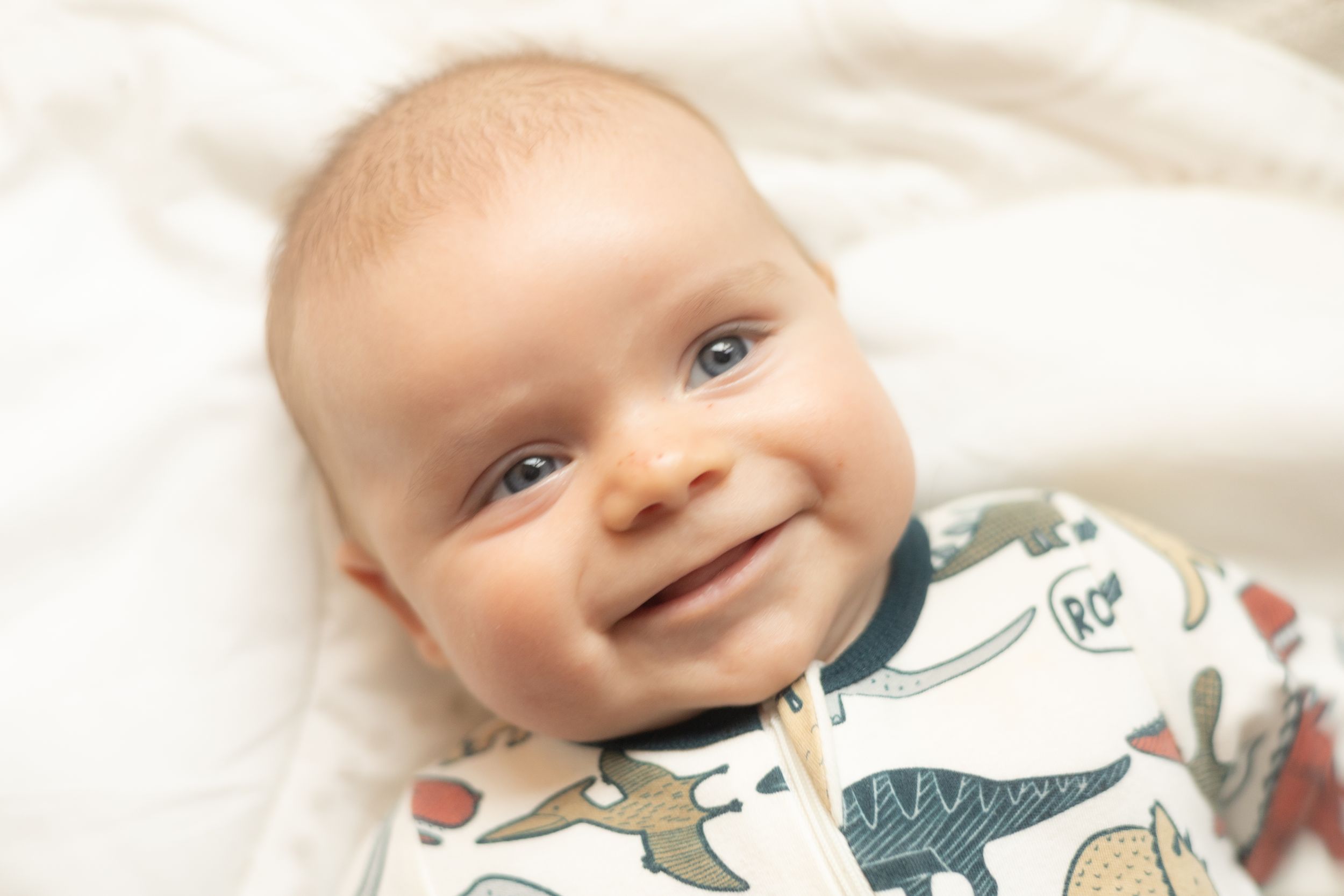 Baby wearing animal print onesie smiles joyfully while lying on white bedding.