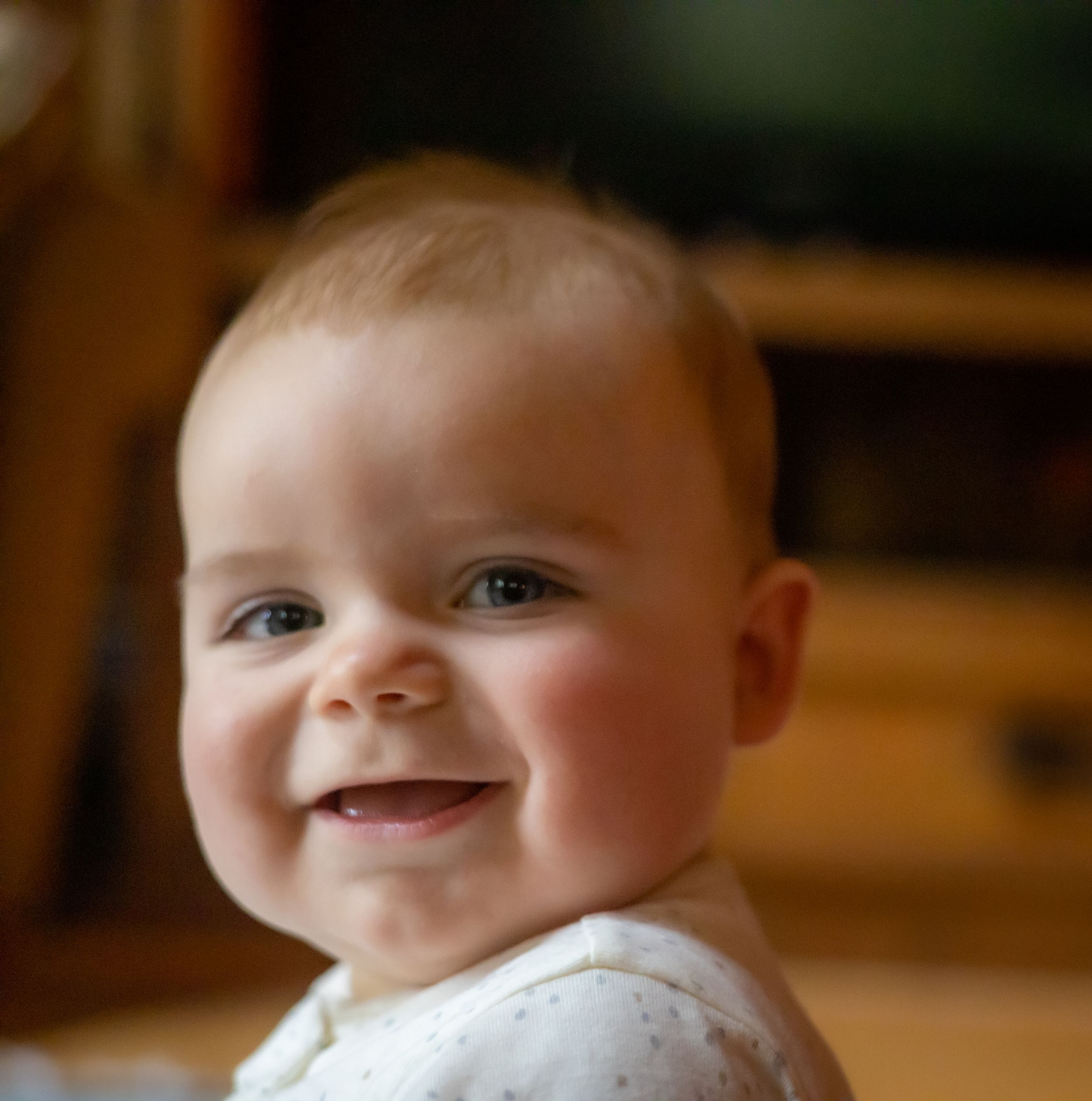 Baby in white outfit smiles brightly at camera with rosy cheeks.