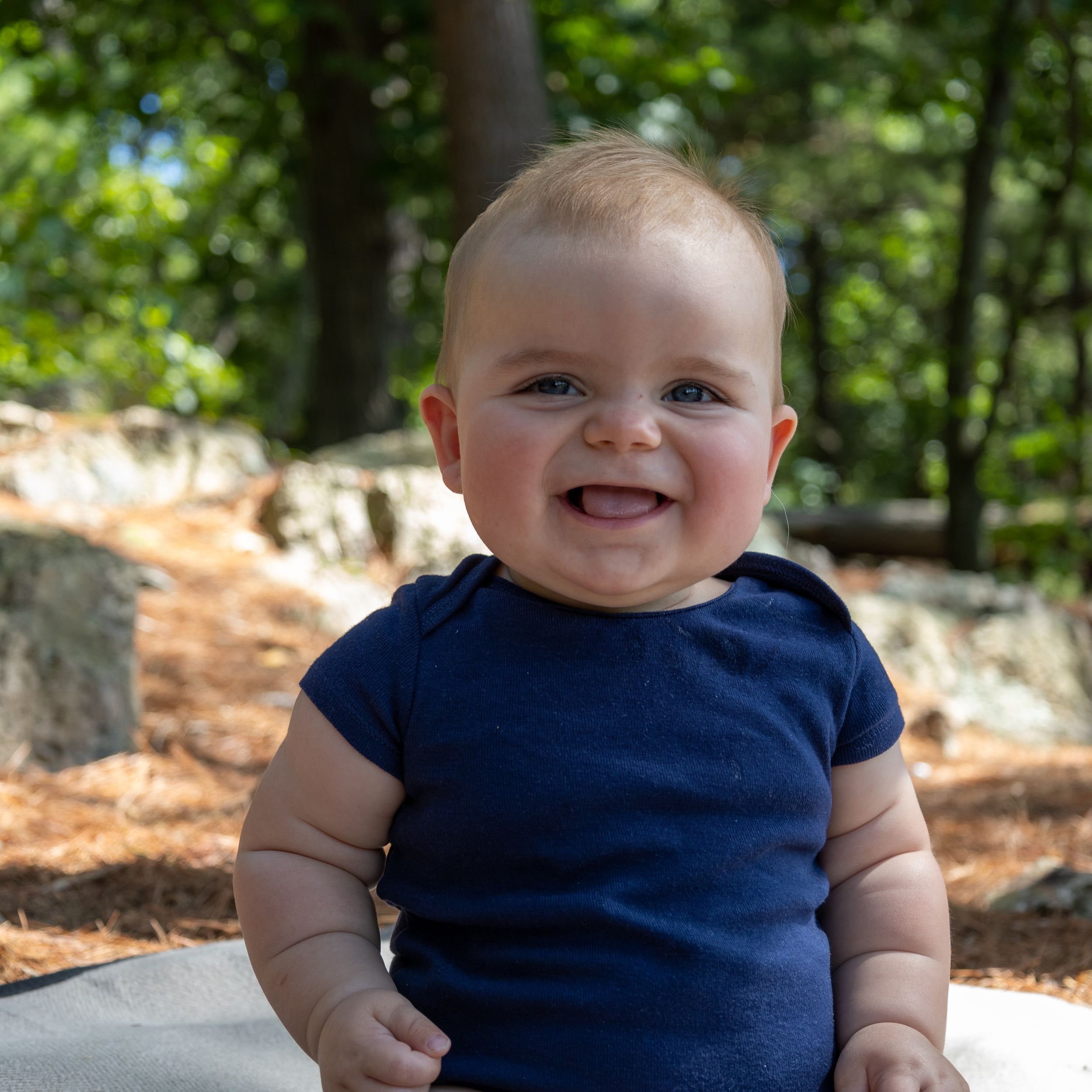 Happy baby in navy blue t-shirt sits outdoors on sunny day with natural background.