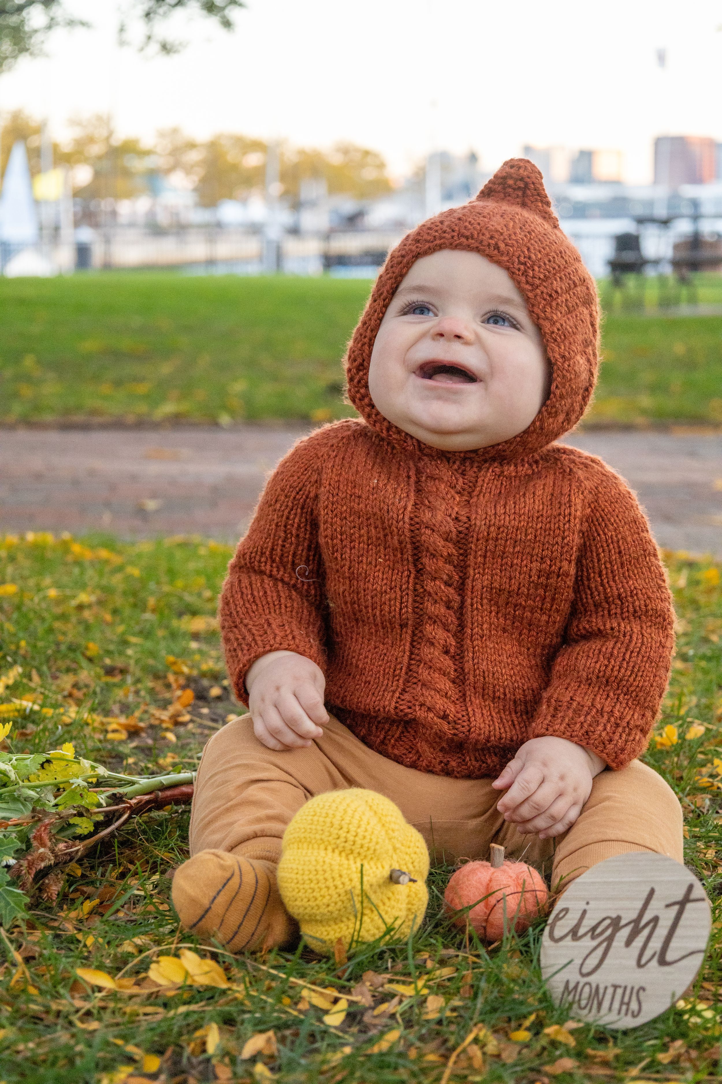 Baby in orange knit hoodie sits on autumn grass with yellow pumpkin decorations.