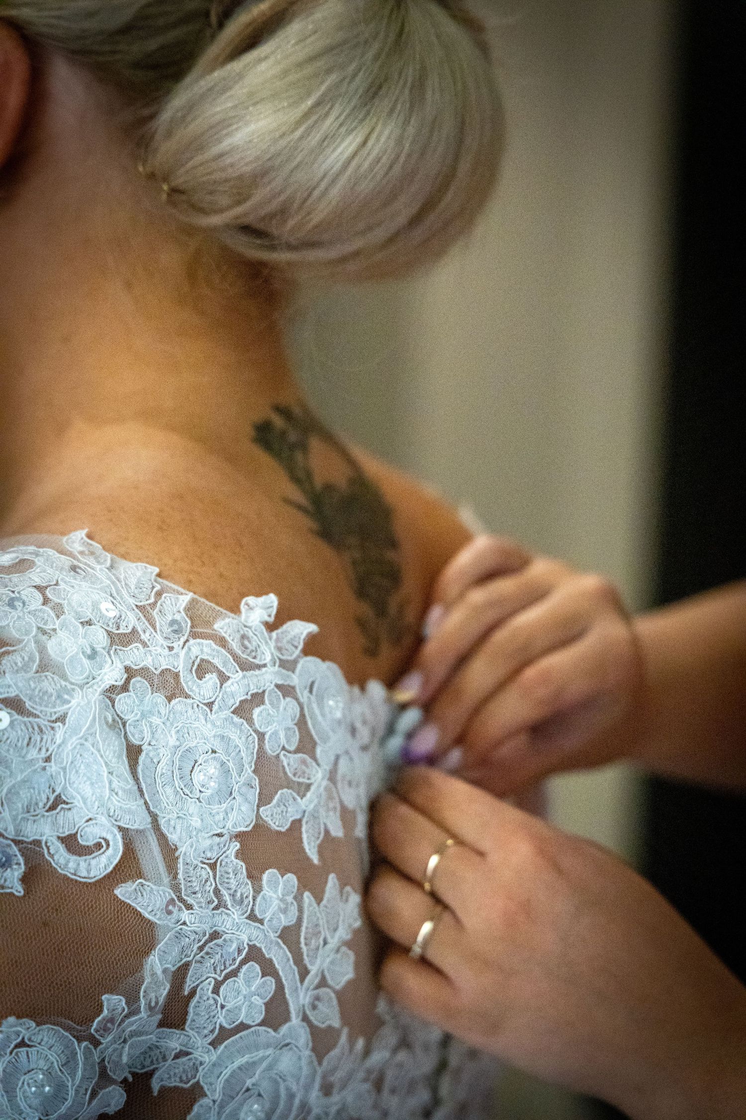 Hands button up intricate lace back of bridal gown on blonde bride with floral tattoo.