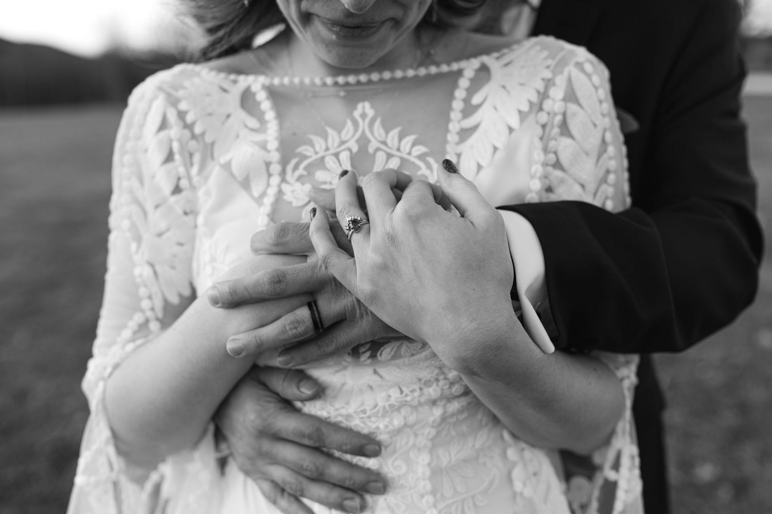 Black and white close-up of hands exchanging wedding rings against a delicate lace dress detail.