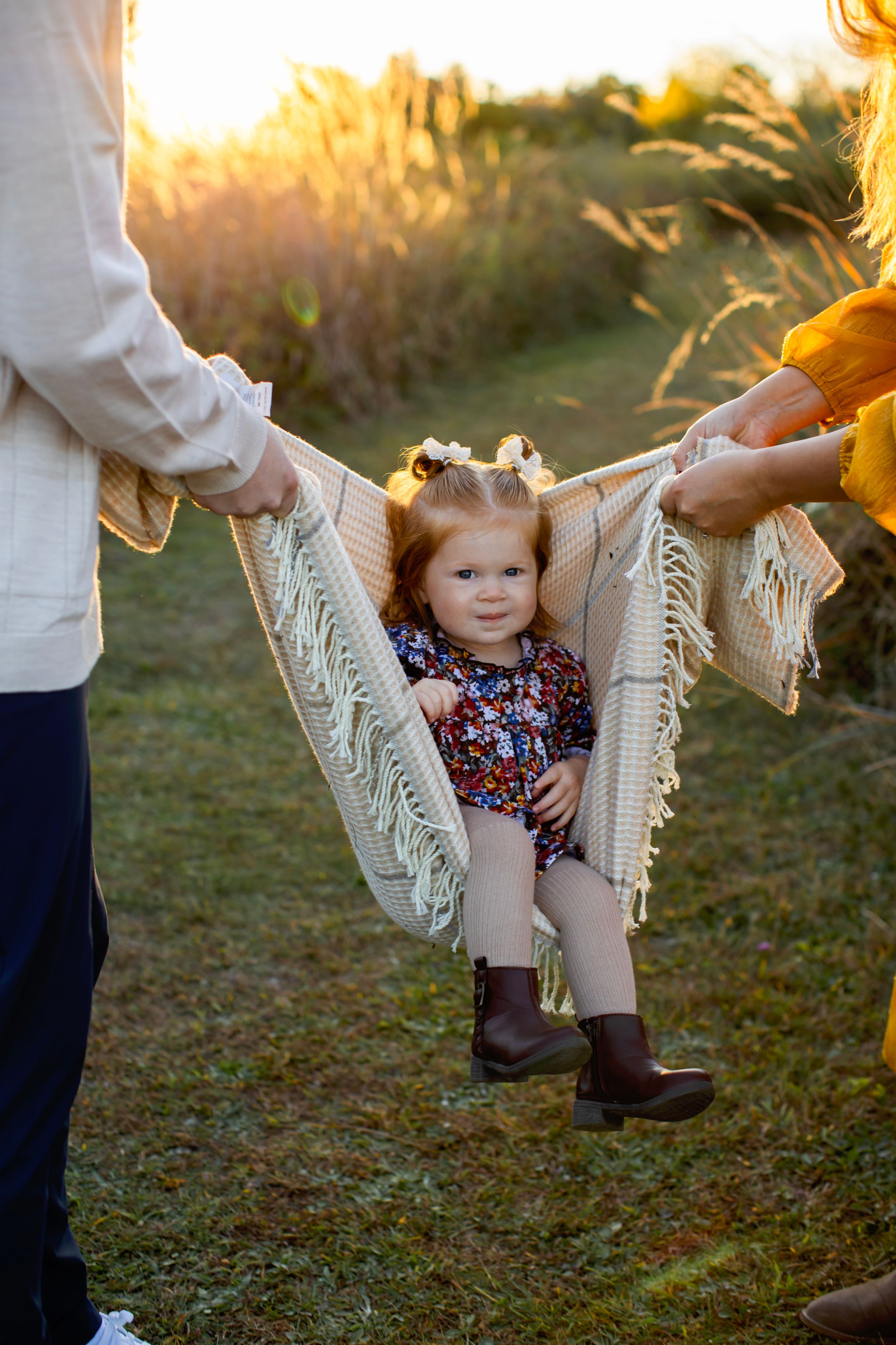 A small child sits in a macrame hammock swing at sunset while being supported by adults on either side. NJ Family Photographer