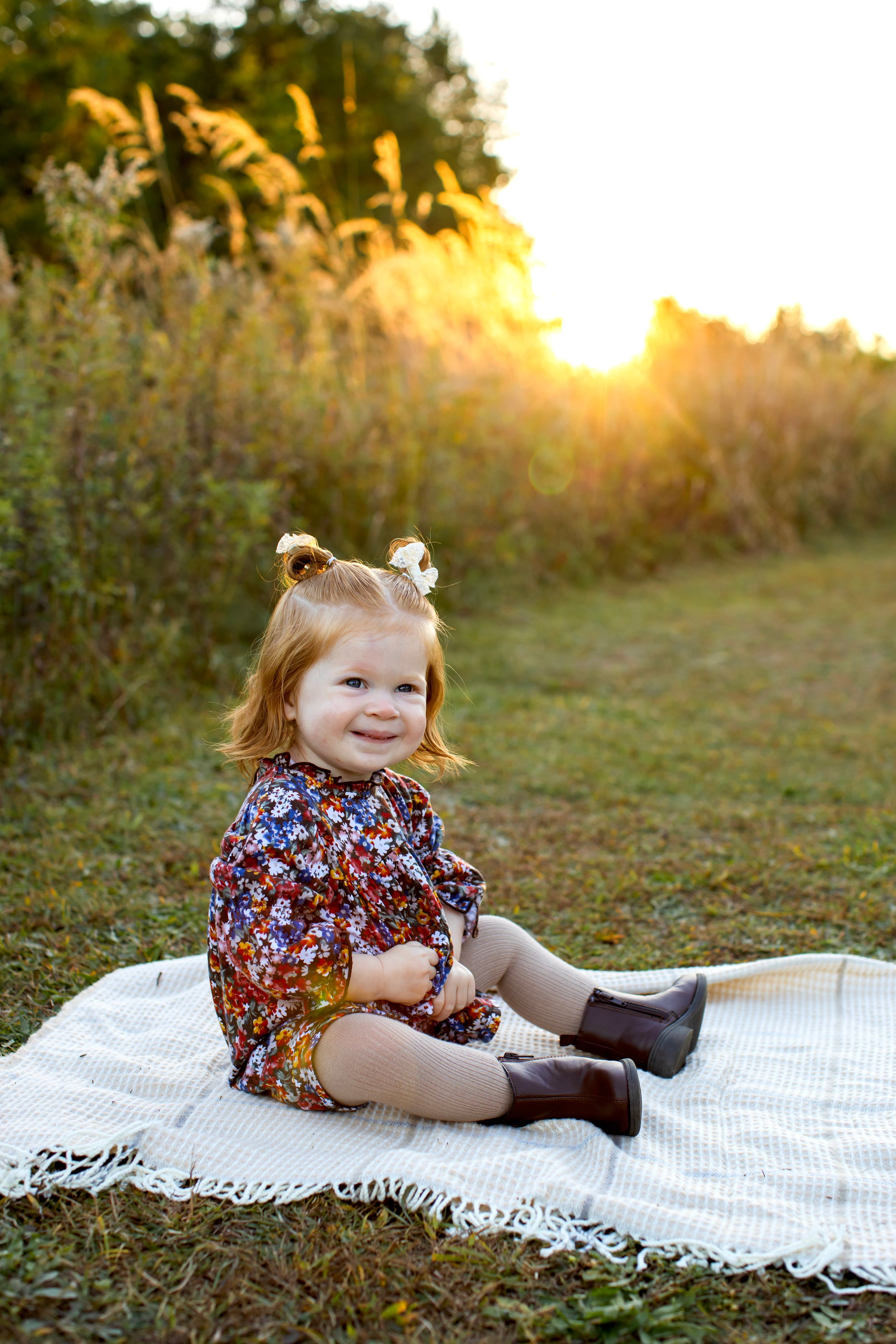 A floral romper and tiny boots on a white blanket during golden hour sunset with tall grasses in the background. NJ Family Photographer