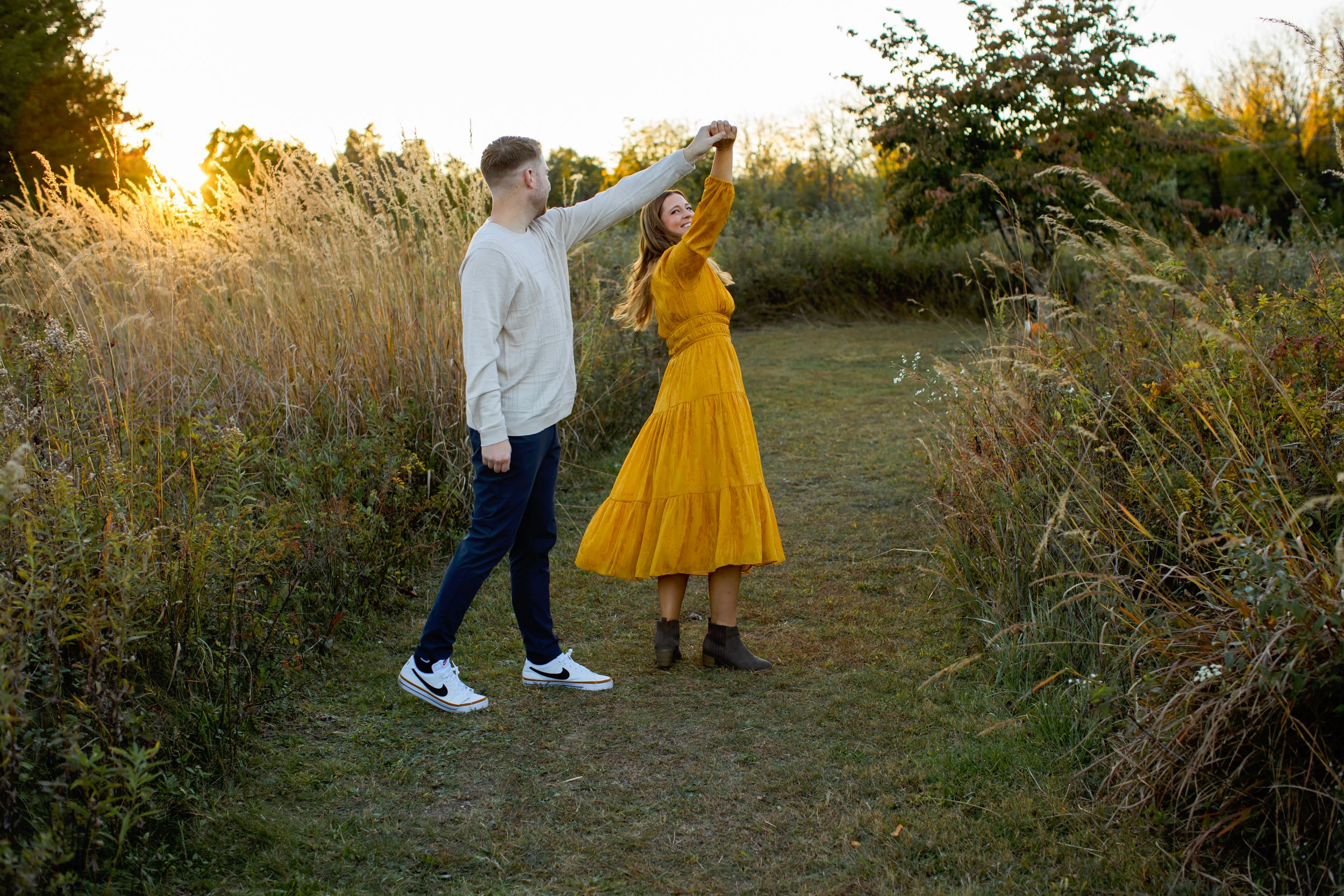 Two people dancing together on a path at sunset, one wearing a vibrant yellow dress and the other in casual attire. NJ Family Photographer