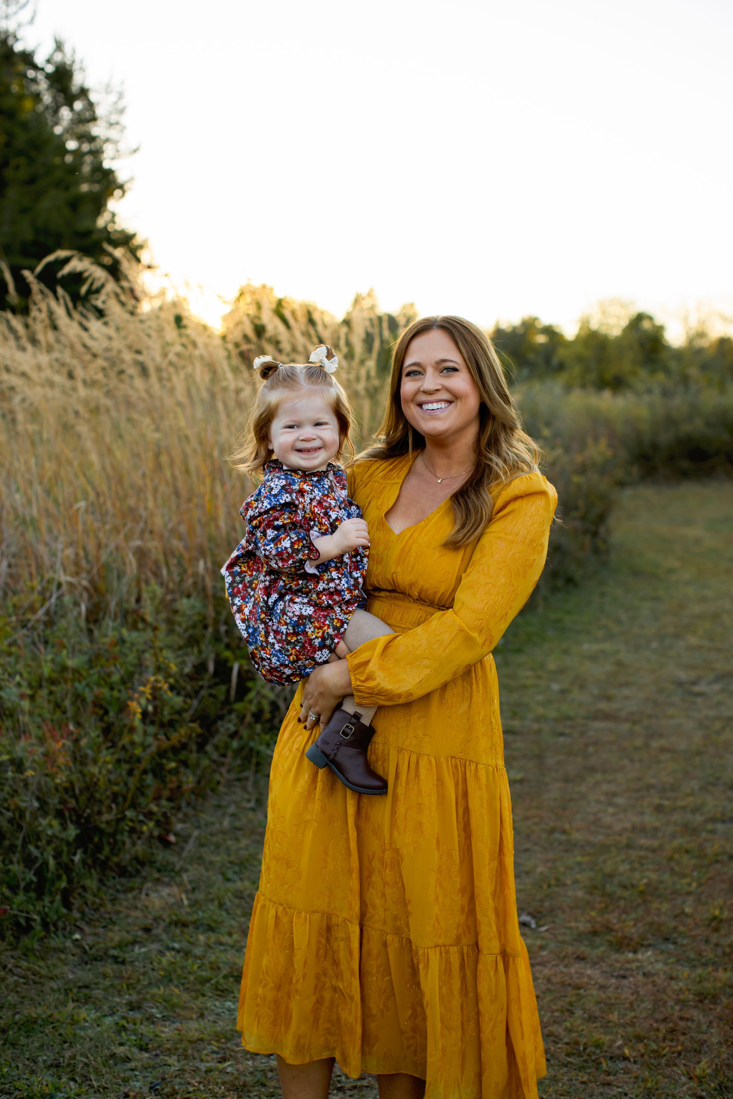 Mother in mustard yellow maxi dress holds small child wearing a floral outfit in golden sunset field. NJ Family Photographer