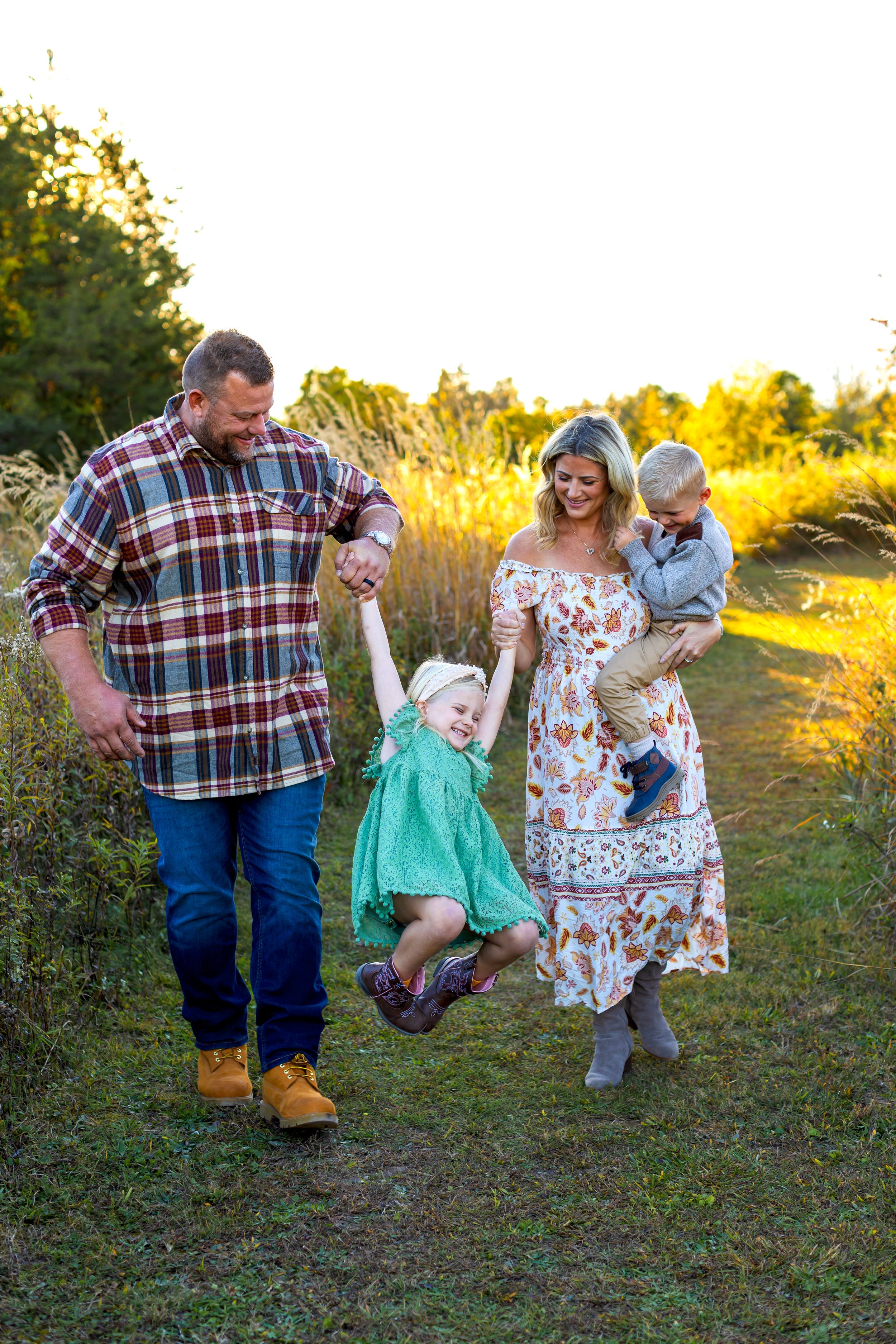 A family plays together outdoors at sunset, with a small child jumping playfully on a grassy path. NJ Family Photographer