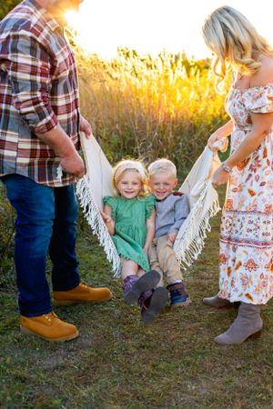 Family poses together in a golden field at sunset with children sitting in a cozy white hammock. NJ Family Photographer