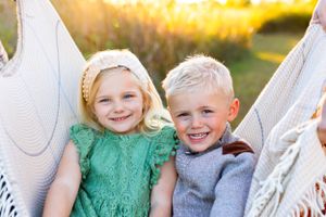 Two young children in a hammock smile brightly at sunset, one wearing green and one in gray. NJ Family Photographer