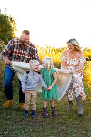 Cheerful family laughing together in a sunset field with tall grass and golden sunlight. NJ Family Photographer