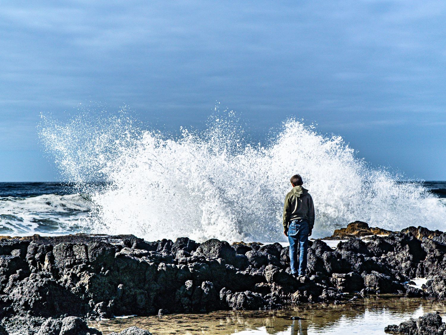 Large ocean wave crashes behind rocky coast creating dramatic splash.