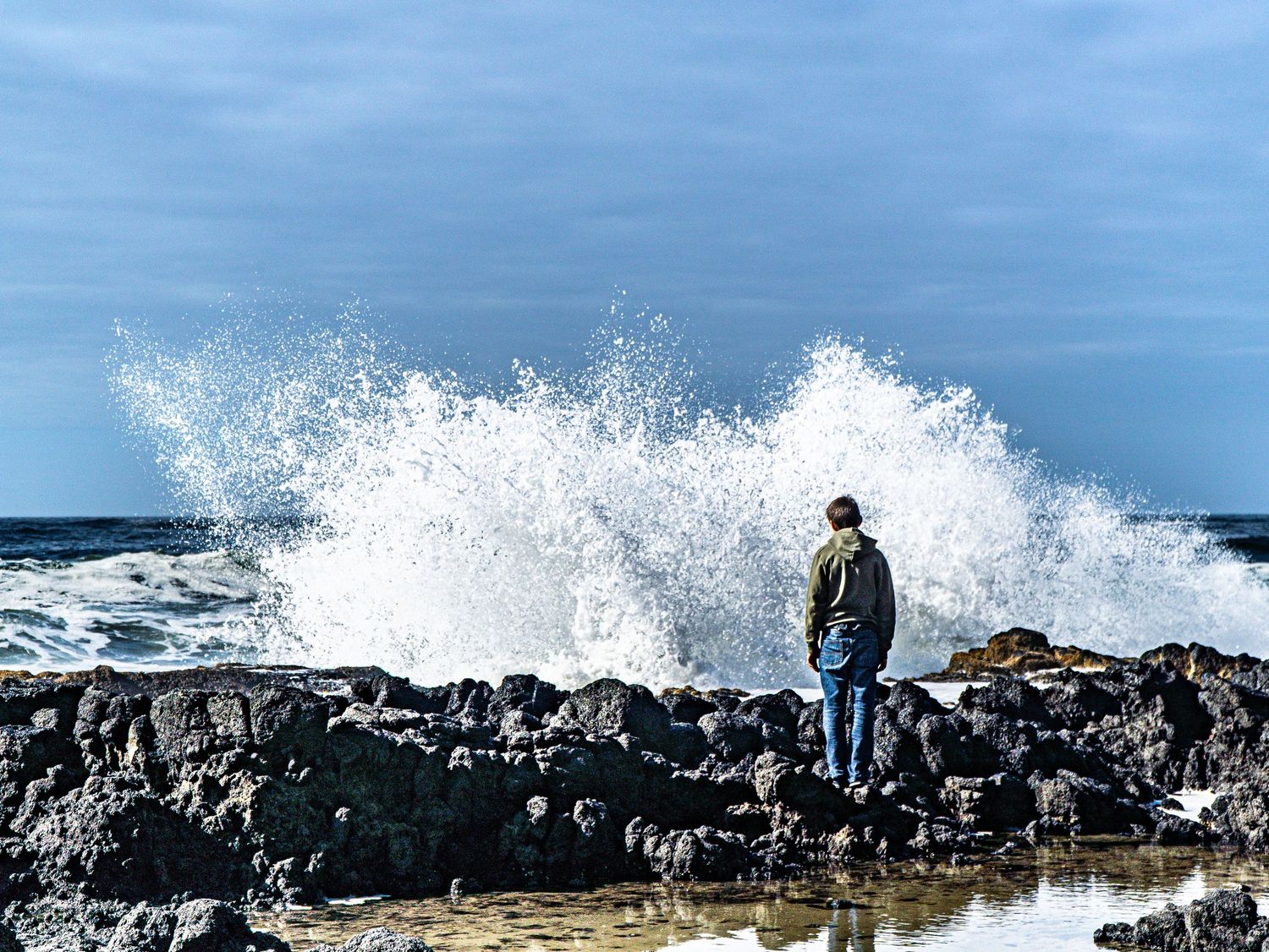 Person standing on rocky shore as large wave crashes dramatically behind them.