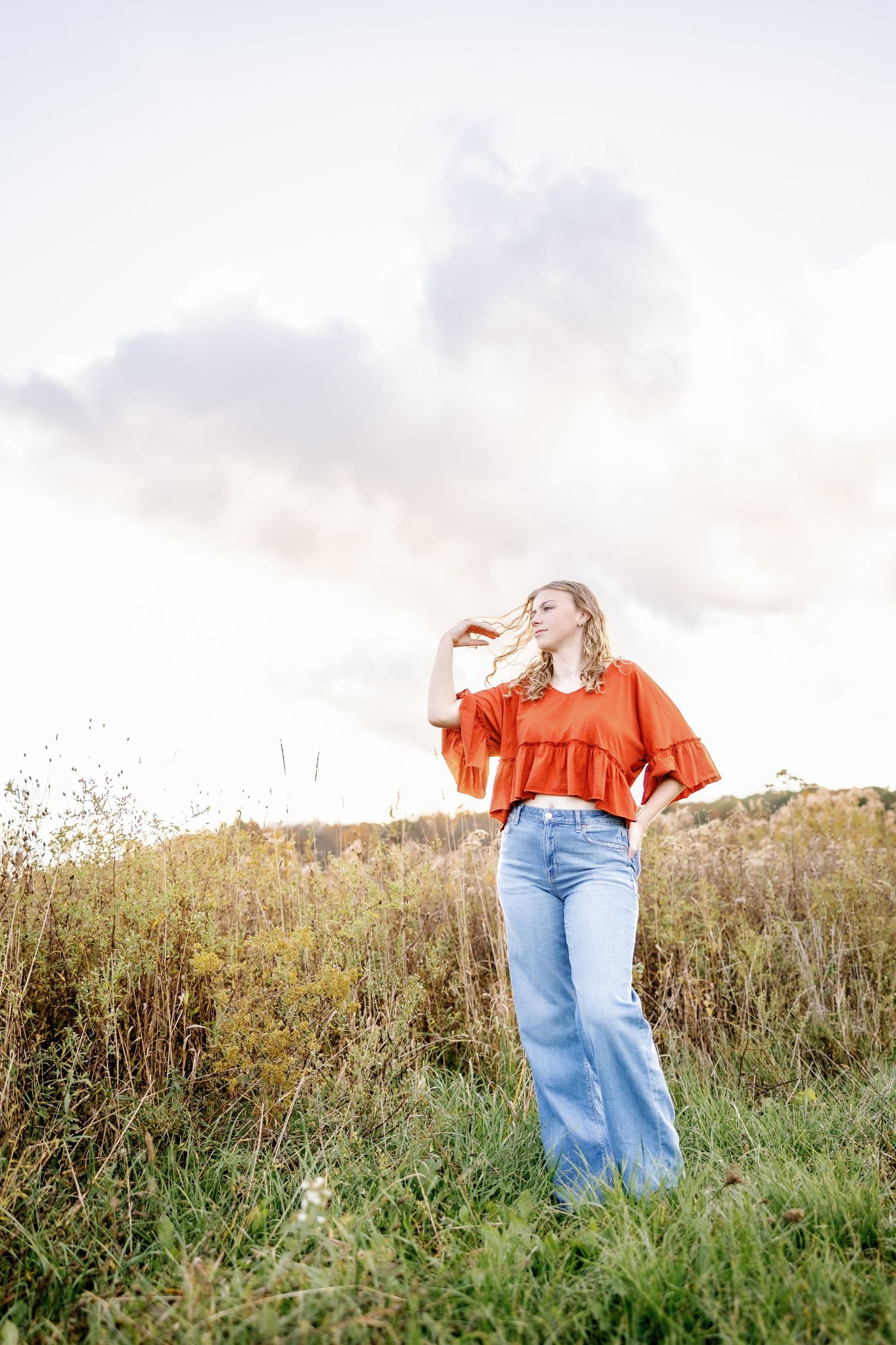 Senior in bright orange top poses playfully in a sunny wildflower meadow at sunset.