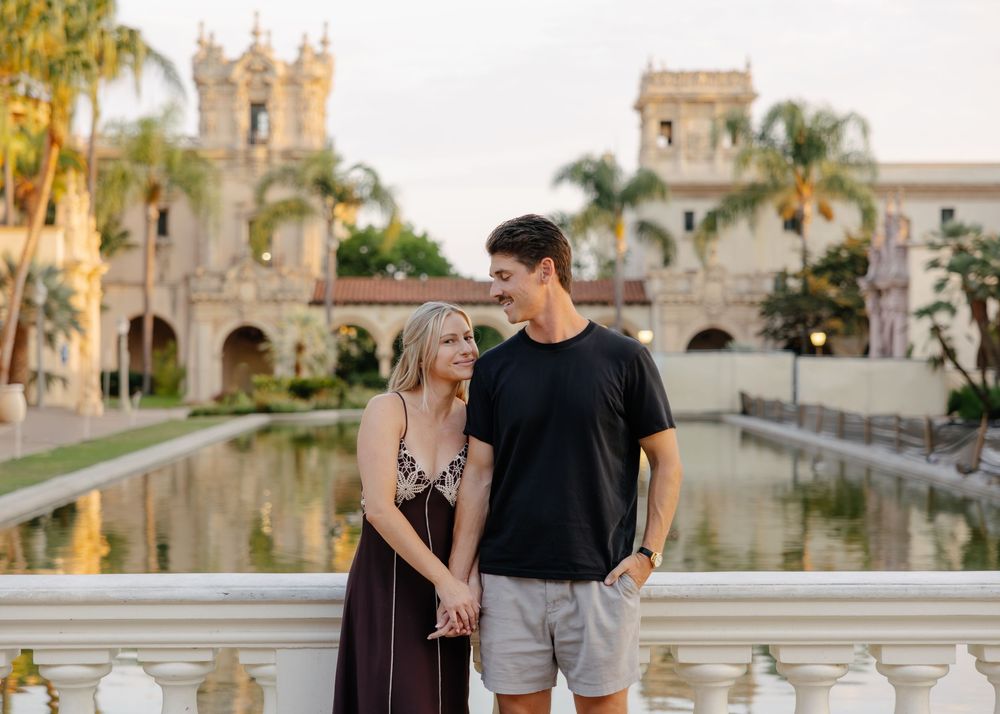Romantic couple strolling along an outdoor plaza with Spanish colonial architecture and palm trees in the background.