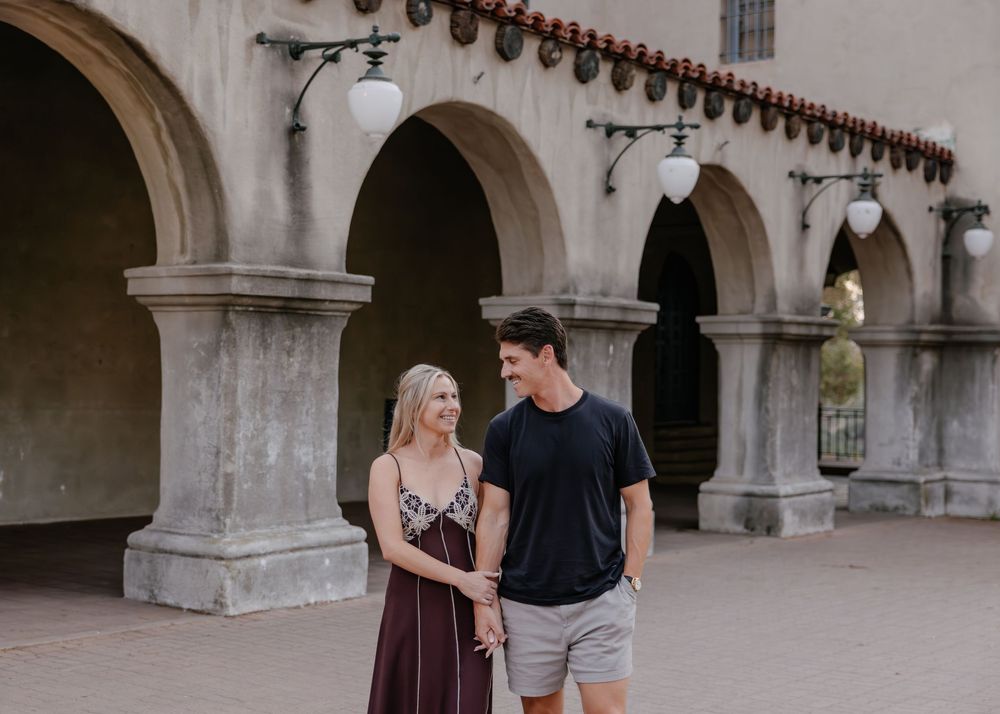 A happy couple interacts under a series of arched columns in a Spanish mission style building.