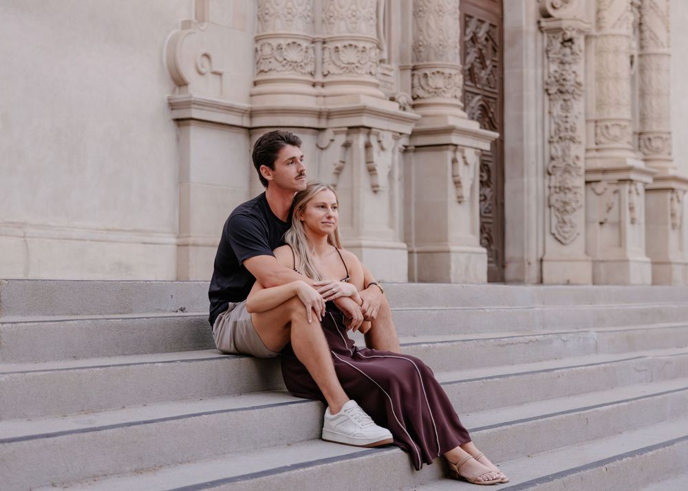 A couple sits together on stone steps of an ornate building with classical architecture.