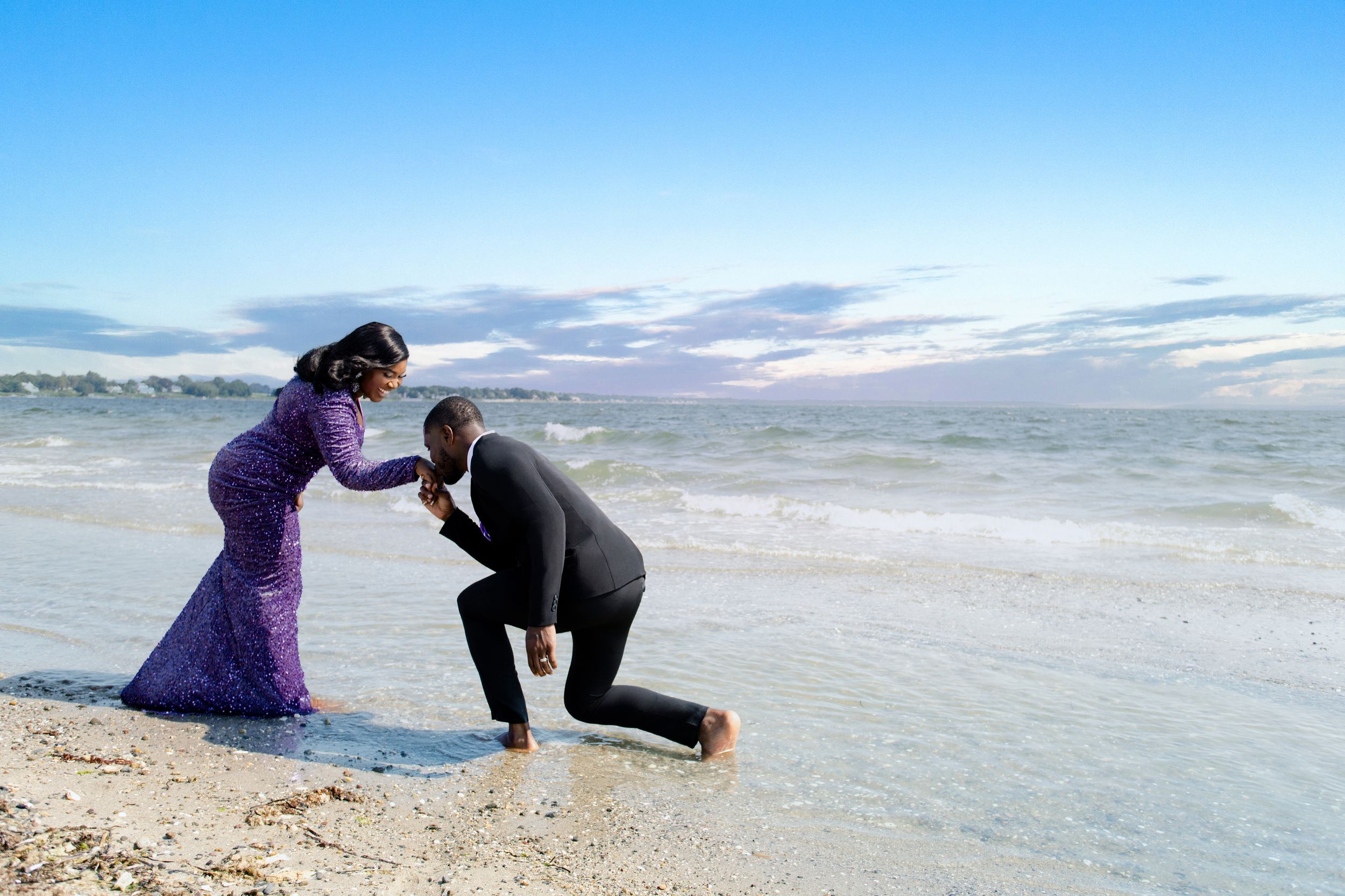 A romantic beach proposal scene with one person kneeling in front of another against a blue sky.