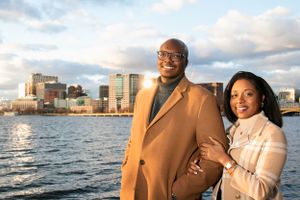 Couple in camel coats pose together with city skyline and water in background.