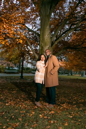 Couple in matching camel coats pose under large autumn tree with orange leaves.