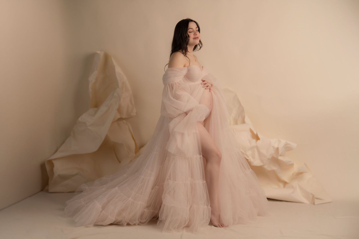 A pregnant woman poses in a flowing blush pink tulle gown against a cream backdrop with billowing fabric. Northeast ohio