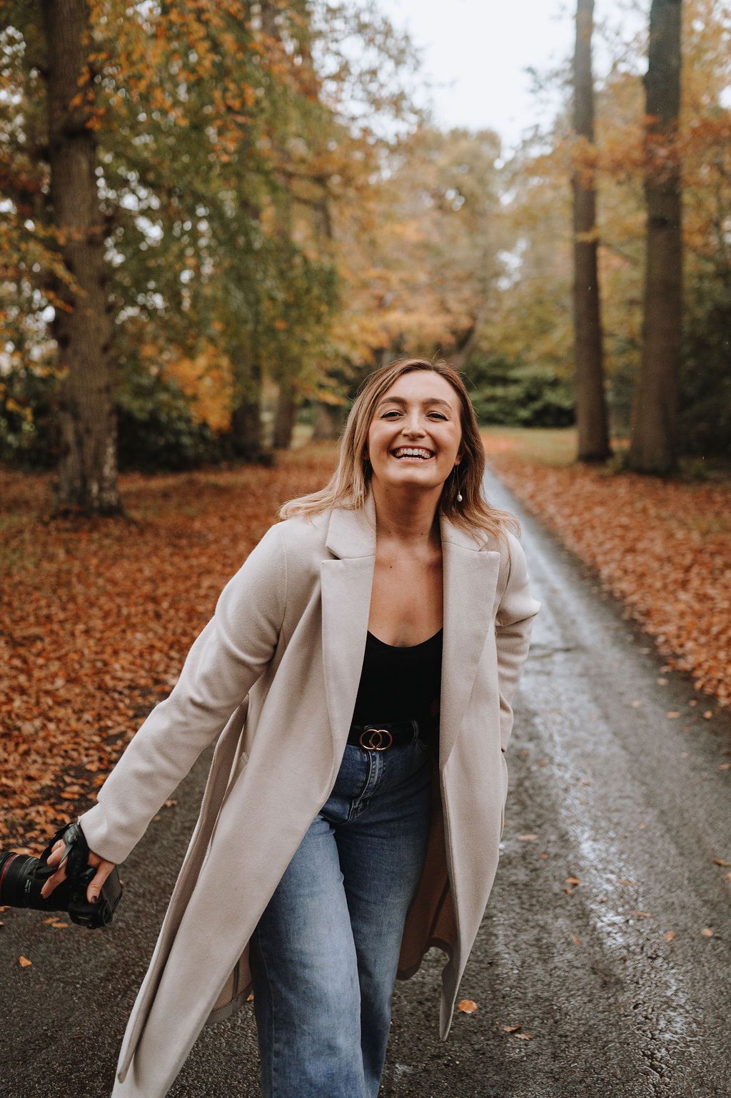 A casual autumn outfit featuring a beige coat and jeans surrounded by fall foliage on a wet path through the woods.