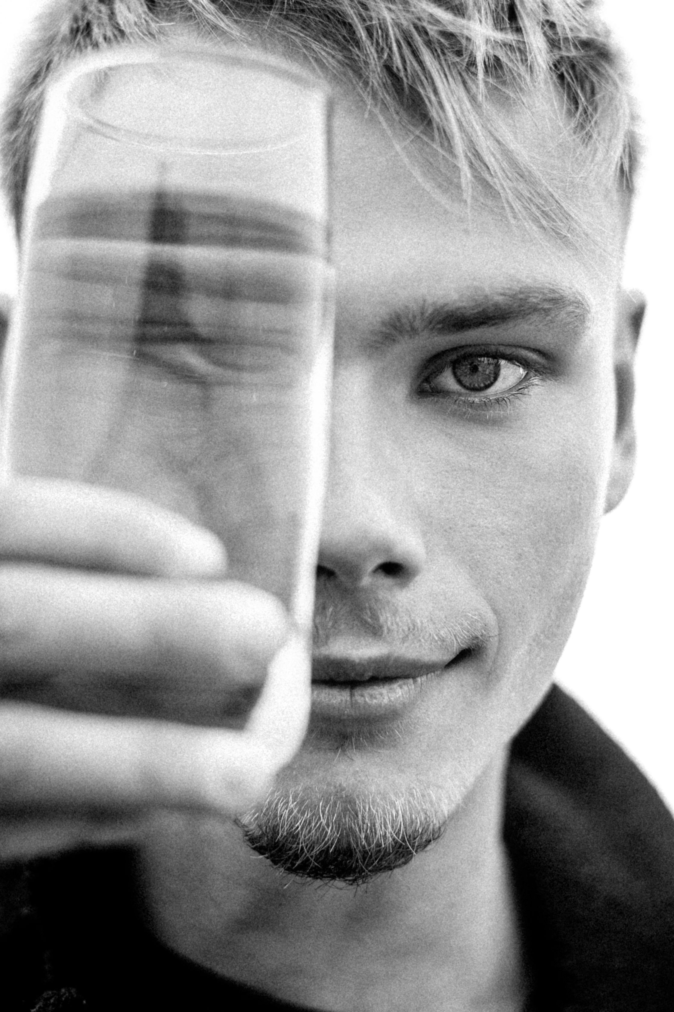 Headshot of a young man holding a glass in front of his face. Black and white portrait of a male model