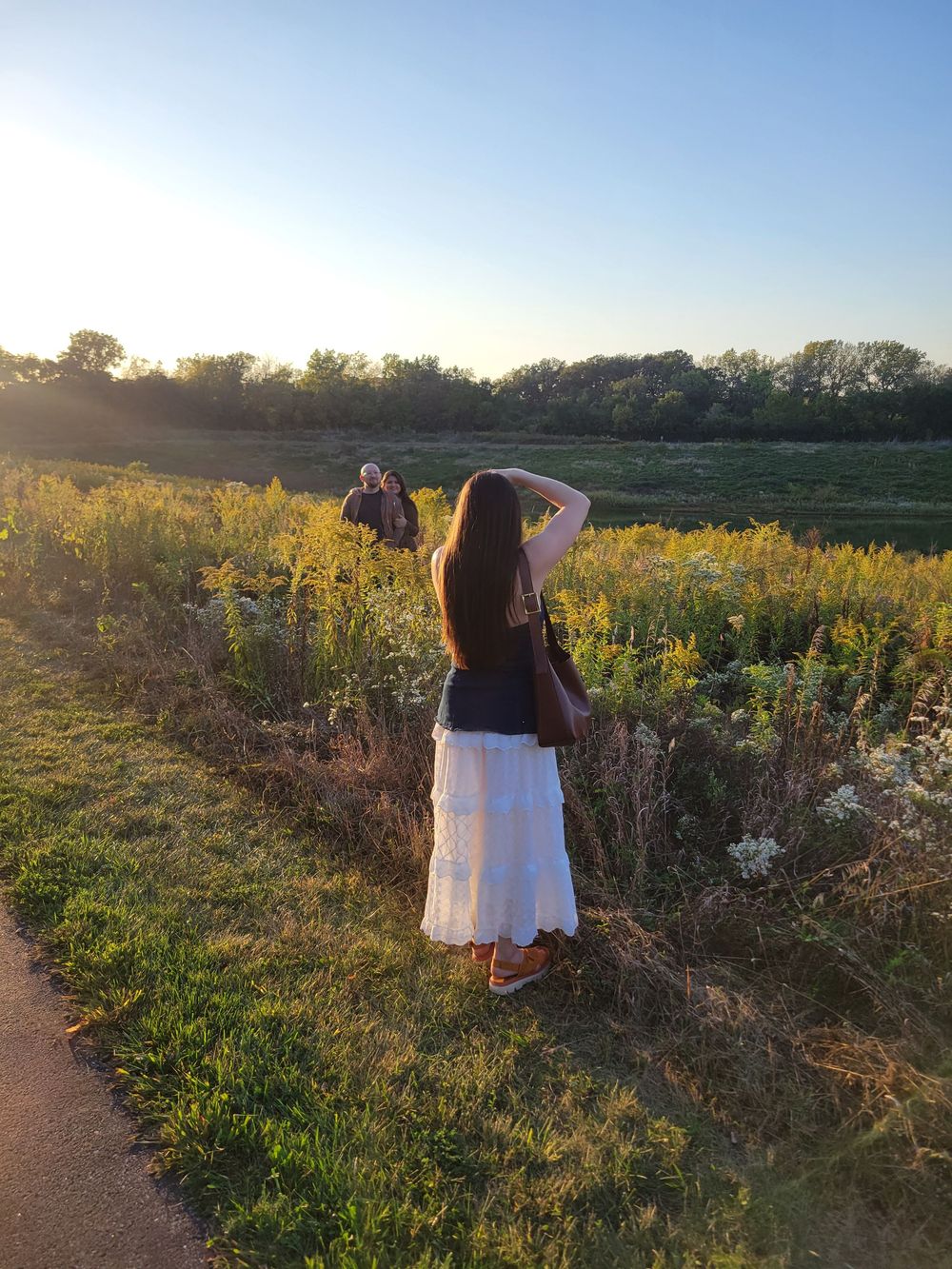 A person in a brown top and white skirt stands in a wildflower meadow at sunset taking photos of the landscape.
