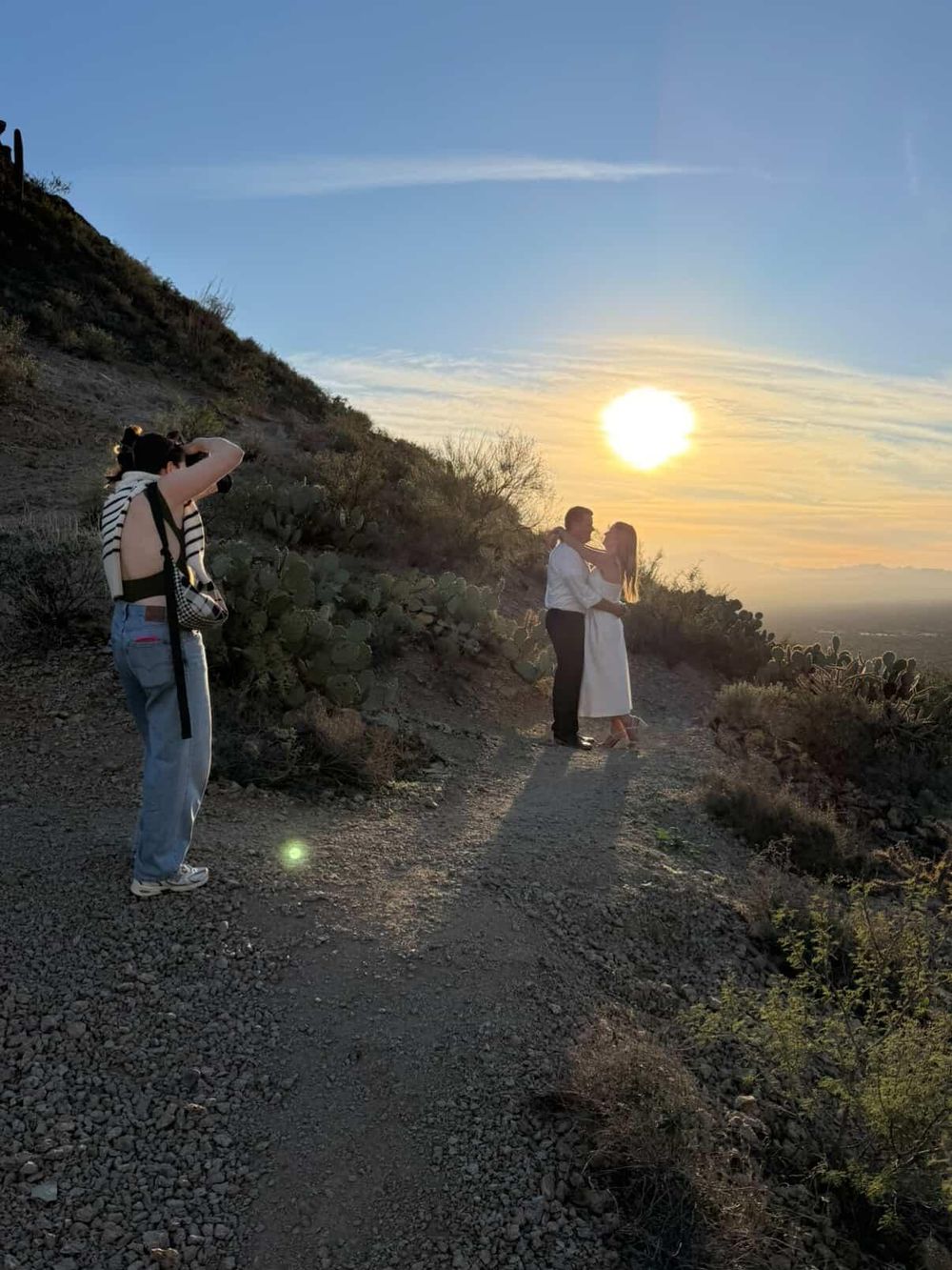 Hikers traverse a desert trail at sunset while capturing photos of the golden sky and mountainous landscape.
