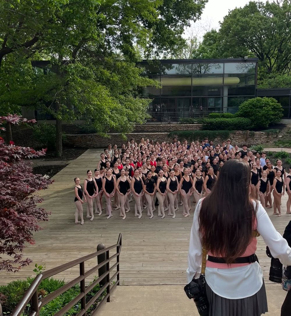 Large group of dancers in black leotards arranged in rows on an outdoor wooden deck during a performance.